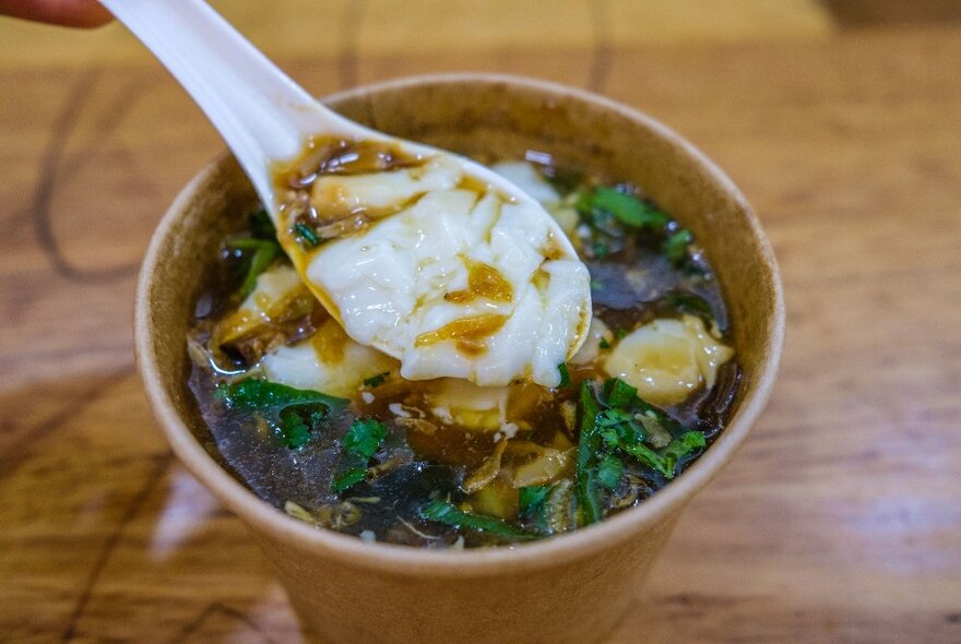 A small white spoon lifting up a thick soup from a takeaway soup bowl.