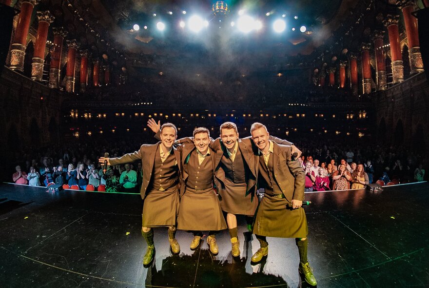 The group Celtic Thunder posing for a photo in a concert hall wearing kilts with the audience visible behind them. 