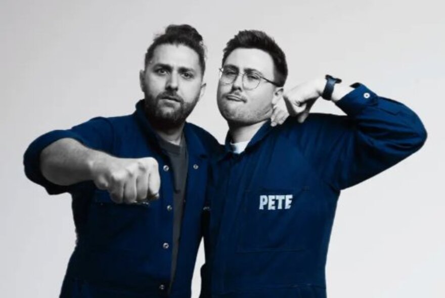 Two men wearing overalls, one pushing his fist forward, the other with his hand to his head.