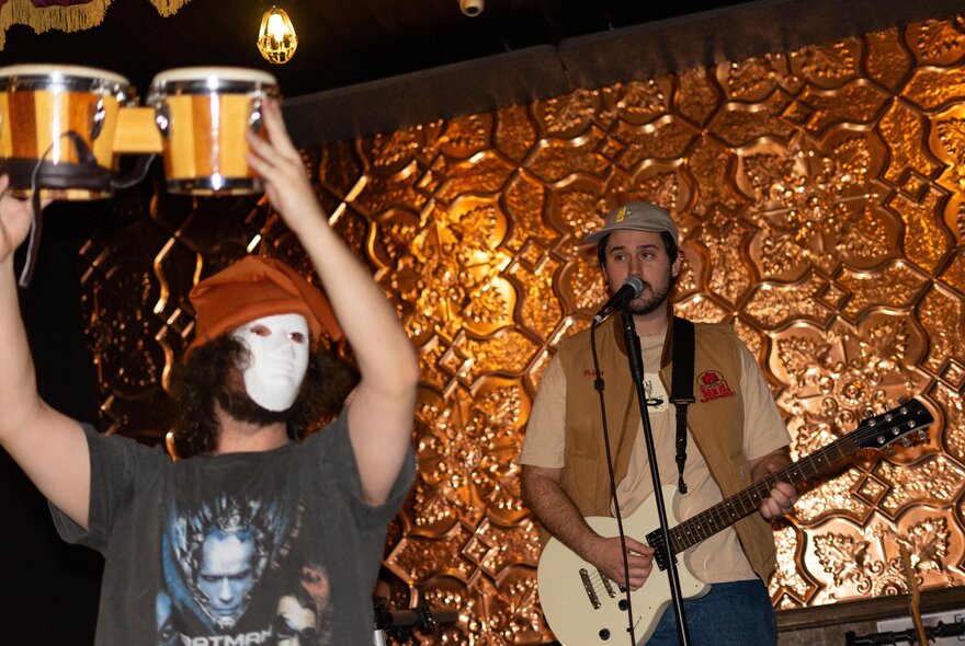 Musicians on stage, one wearing a white mask and holding up a set of bongos while the other plays the guitar and sings against an ornate gold background. 