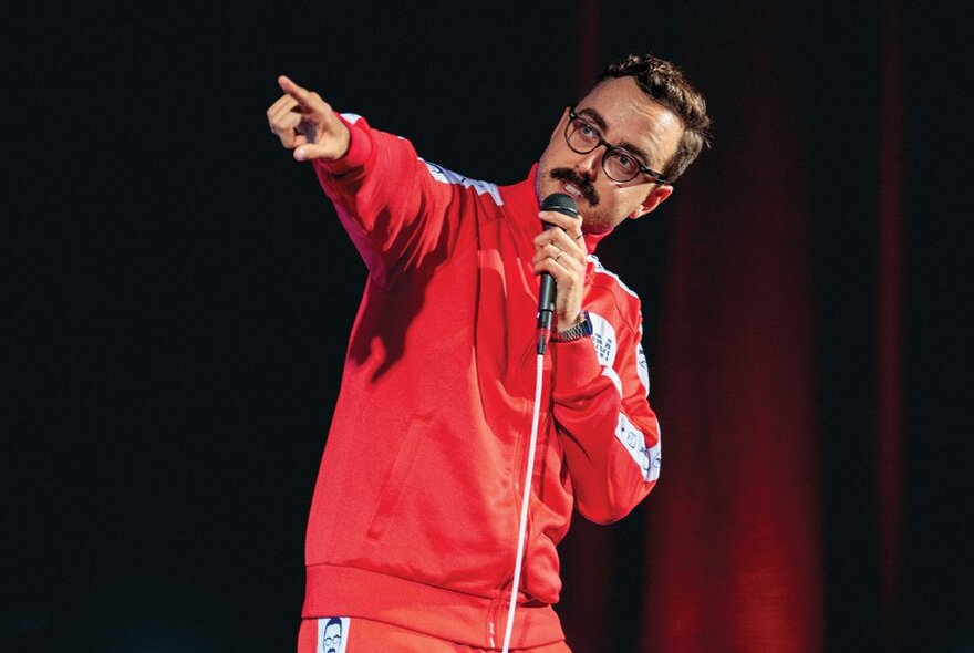 A man wearing a red tracksuit and holding a microphone that is on a stand, performing a stand-up routine on stage and pointing with one arm.