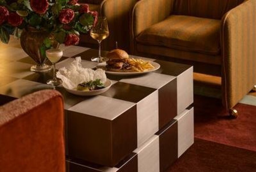 A lounge area with a brown and white checkered coffee table, two upholstered chairs, a bouquet of red roses in a vase, and a plate with a burger and fries.