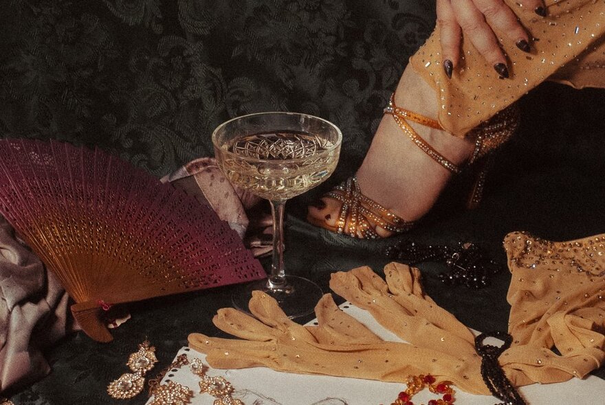 Gloves and a champagne glass on a table next to a gold-sandalled foot.