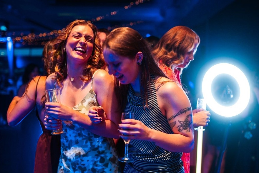 A lively group of women laughing and dancing together under blue and ring-light illumination at an evening social event.