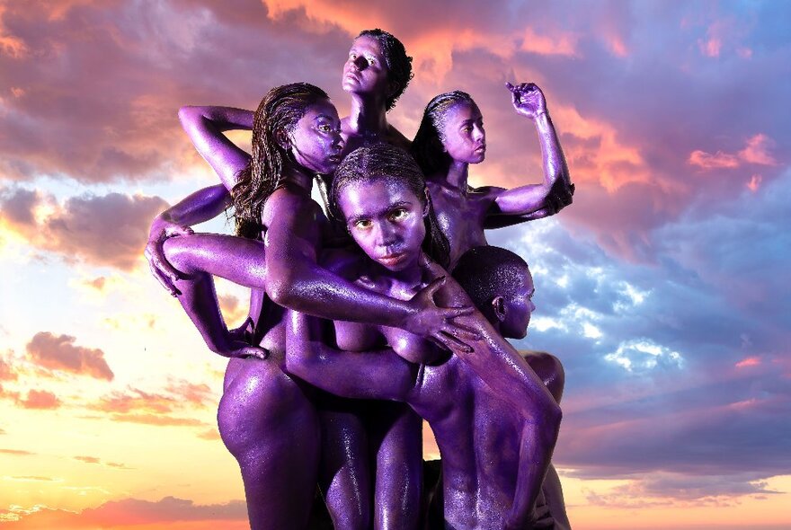 Five naked Brown women entangled together in an impossible embrace, a cloudy sunset sky in the background.