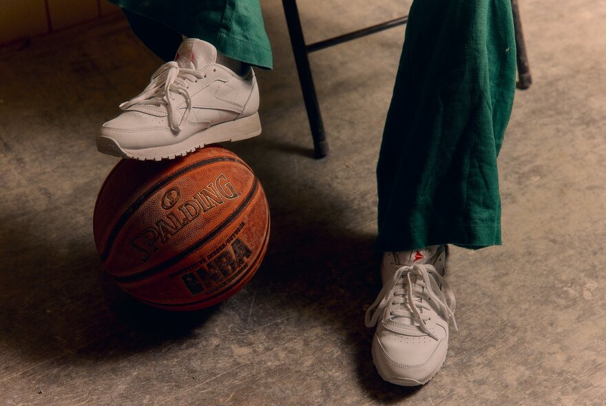 Two feet in white runners, one resting on an orange basketball.