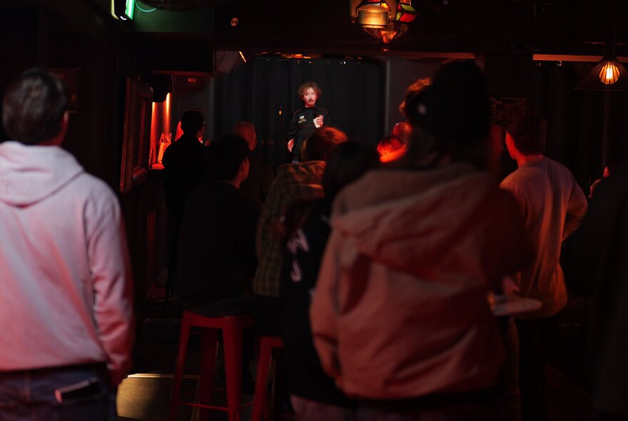 Intimate, very dimly lit indoor club, with a stand-up comedy performer on stage and a seated audience in the foreground.