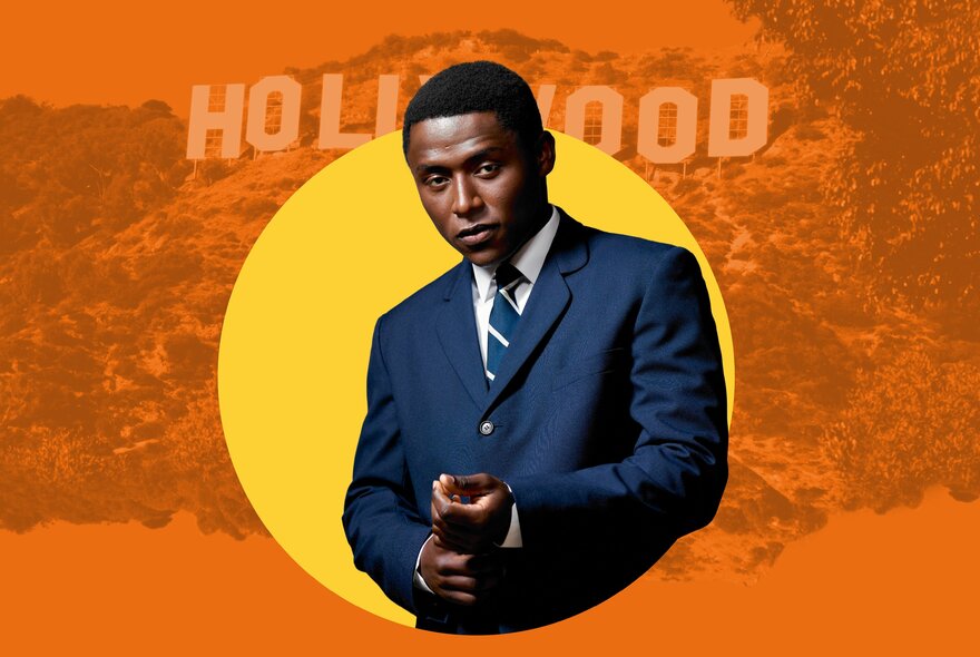 A Black man in a dark blue suit and tie, featured against an orange and yellow background, looking directly at the camera with a serious expression with the word 'HOLLYWOOD' faintly visible in the background. 