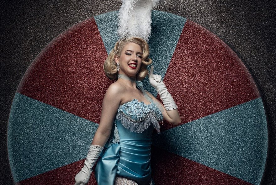 Burlesque performer wearing feathered headdress and embroidered showgirl outfit with long white gloves, standing inside a target design.