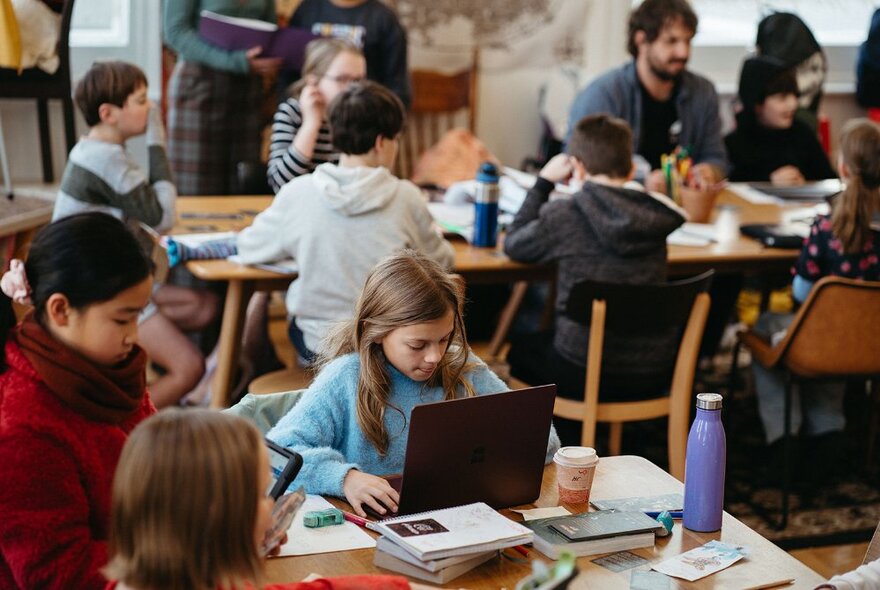 Teenagers sitting and working around  large tables in a room, some with laptops open in front of them, in a bright airy space that is a writer's studio.