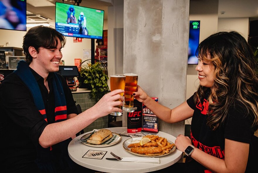 A bar setting with two people clinking their beer glasses over two plates of food, TV on in the background. 