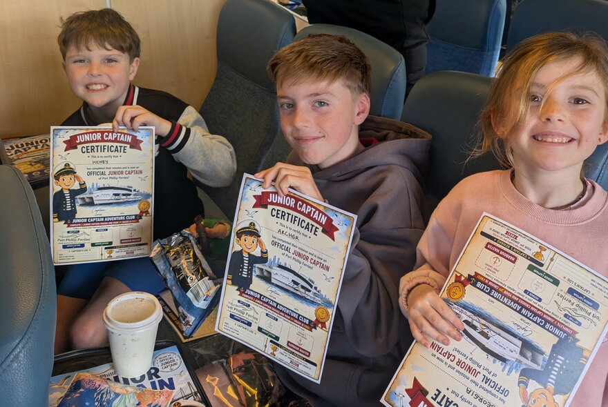 Three young children sitting indoors on ferry seats, smiling and holding up certificates that look like 'Junior Captain' awards.