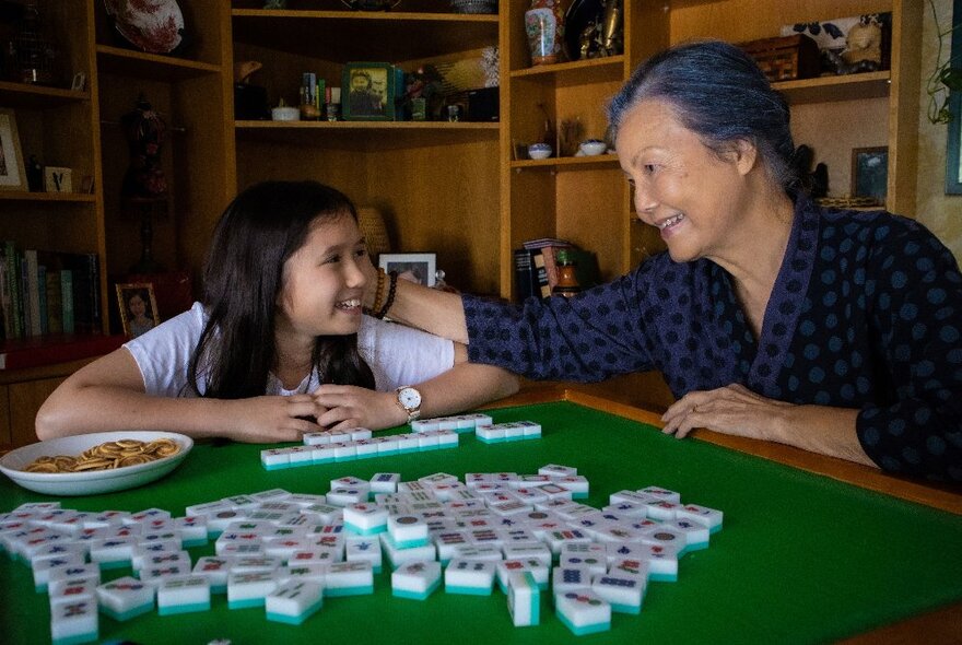 Child and older woman playing mahjong on a green table.