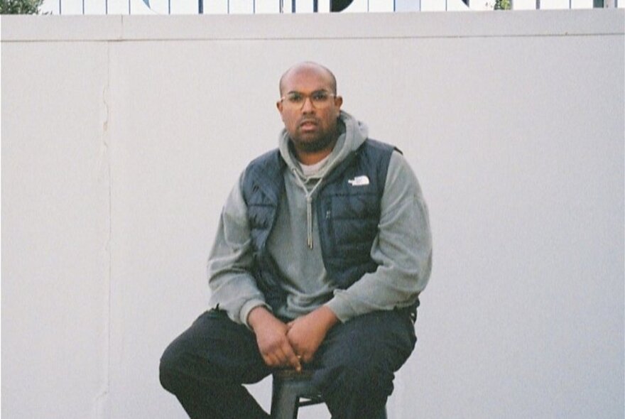 Performer Rowan Thambar wearing jeans and a hoodie, sitting on a stool in front of a white wall.