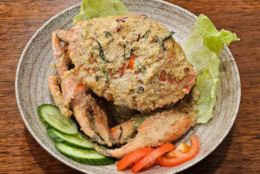 A whole fried crab in a bowl with a few pieces of lettuce, sliced cucumber and tomato.