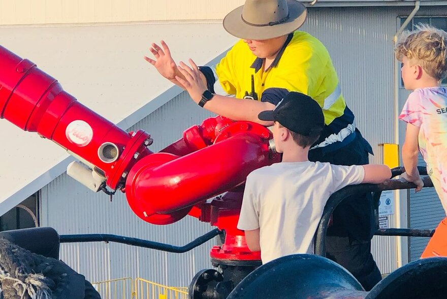 An adult talking to two children in front of a bright red high-pressure fire monitor, or fire water cannon.
