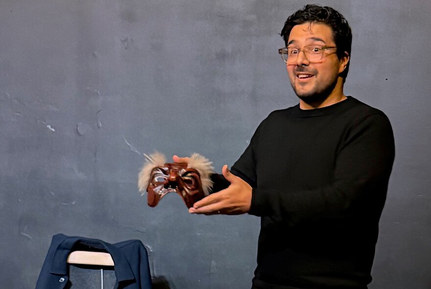 A man in a black long sleeved top holding up a Commedia dell’Arte mask.