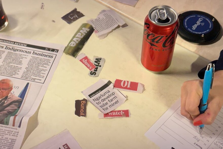 Hand holding a pen completing a form, surrounded by newspaper snippets and a drink can.