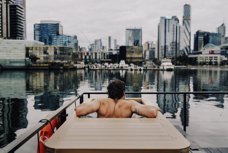 A muscled man seated in a floating sauna moored in Docklands harbour with city buildings in the background.