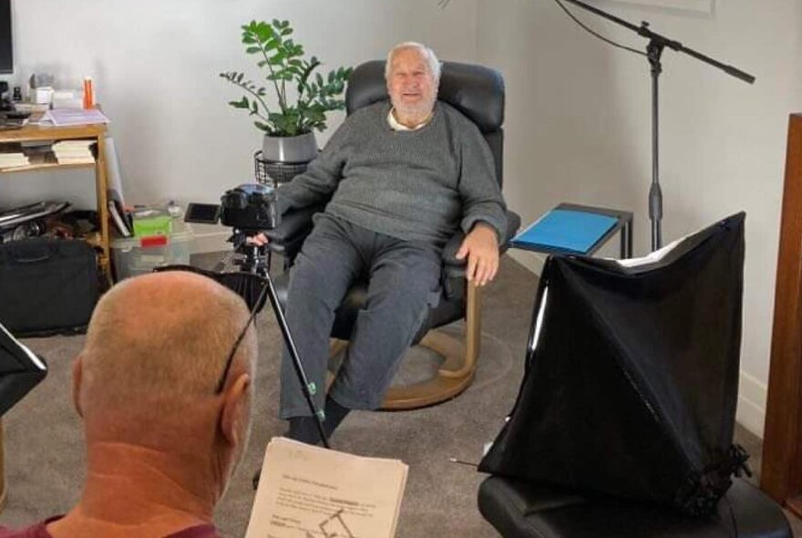 An elderly man seated in an armchair in his lounge room being interviewed and filmed.