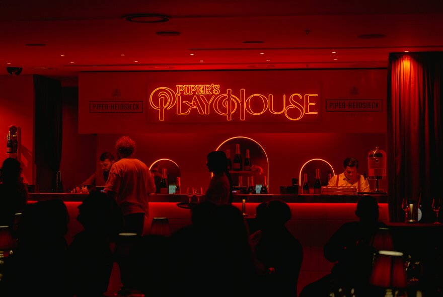 A dark, intimate venue with red lighting and a Piper's Playhouse sign behind the bar. 