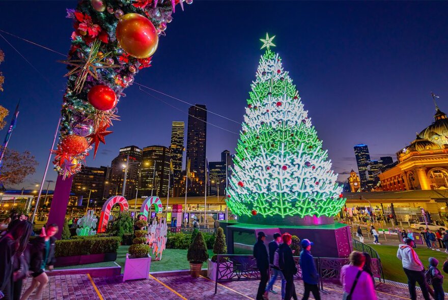 People gathered around an illuminated Christmas tree and decorations at night, Flinders Street Station and city buildings in the background.