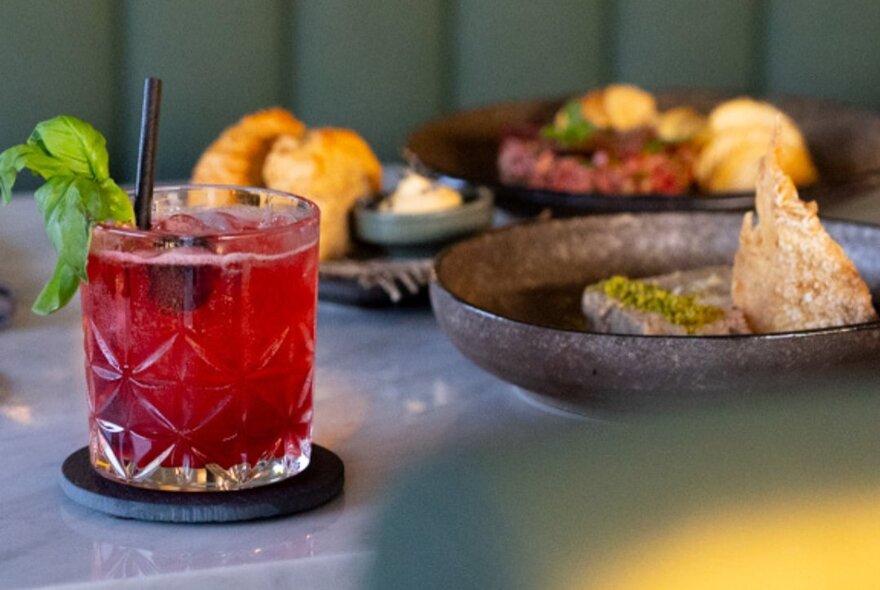 A dining room table with plates of food and a red cocktail on a coaster.