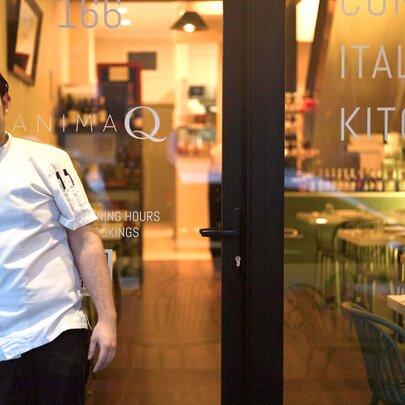 A man in a chef's white shirt standing out the front of a restaurant with large glass windows giving a glimpse of the cosy warmly-lit interior space.