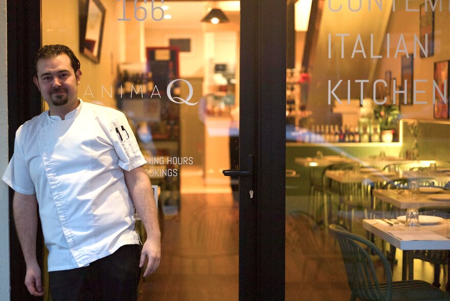 A man in a chef's white shirt standing out the front of a restaurant with large glass windows giving a glimpse of the cosy warmly-lit interior space.