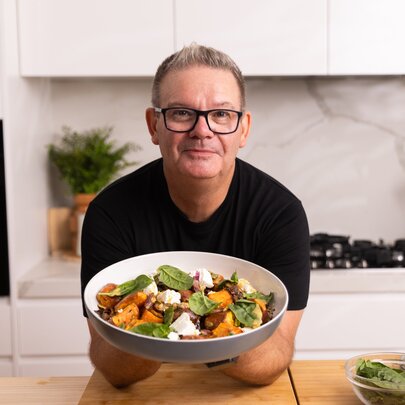 Famous chef, Gary Mehigan, leaning on a wooden bench in a white kitchen and holding up a large bowl of pumpkin and spinach salad.