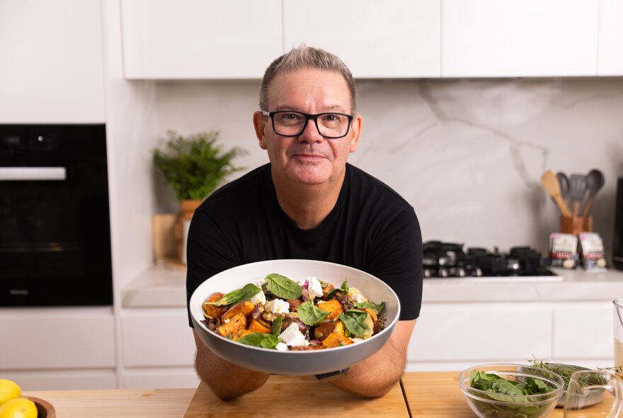 Famous chef, Gary Mehigan, leaning on a wooden bench in a white kitchen and holding up a large bowl of pumpkin and spinach salad.