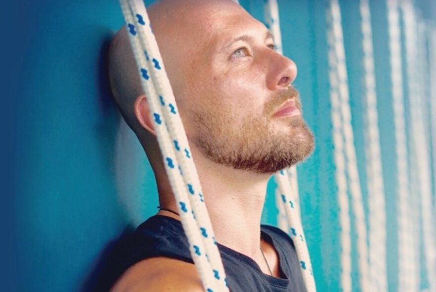 Bearded man standing against a blue backdrop with ropes.