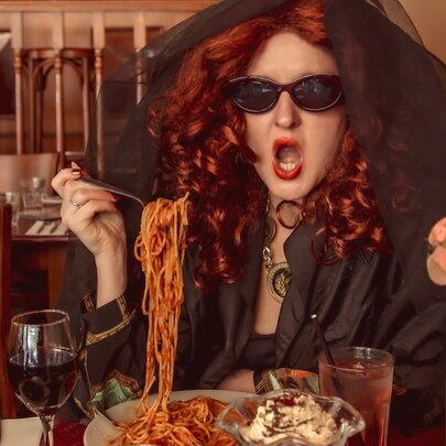 A woman with dark glasses, long red curly hair and a cape hood over her head sitting in a restaurant corner lifting a fork of spaghetti to her mouth. 