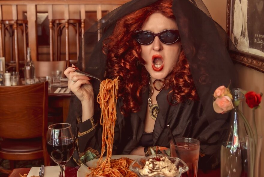A woman with dark glasses, long red curly hair and a cape hood over her head sitting in a restaurant corner lifting a fork of spaghetti to her mouth. 