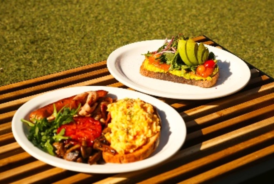 Two plates of breakfast dishes - one with eggs, ones with avocado - in bright sunlight on a wooden striped board on fake grass. 