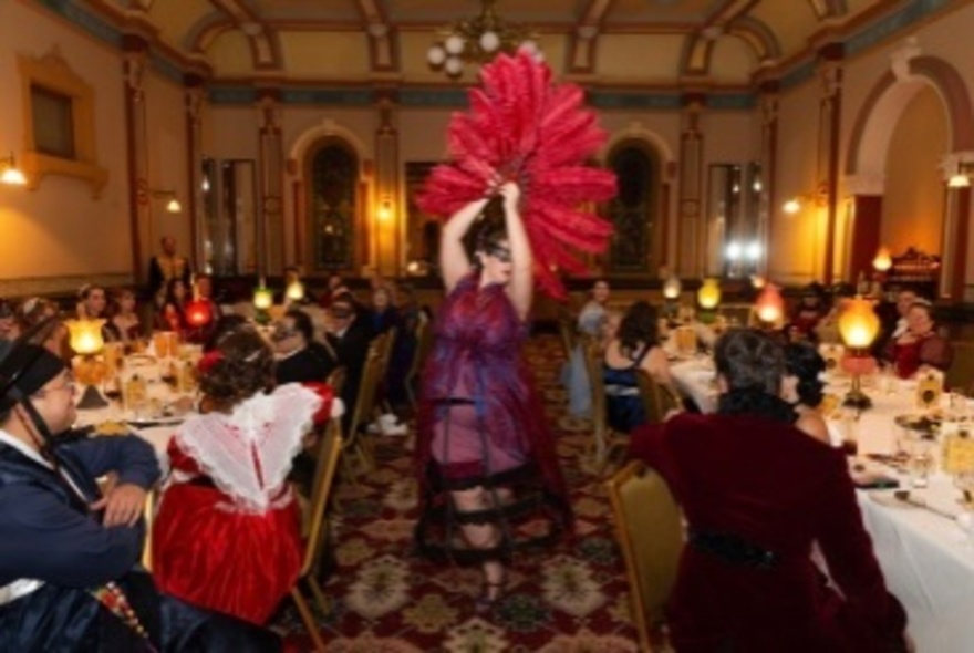 A dancer with a large red feather fan, dancing between seated diners dressed in period costumes in an ornate ballroom. 