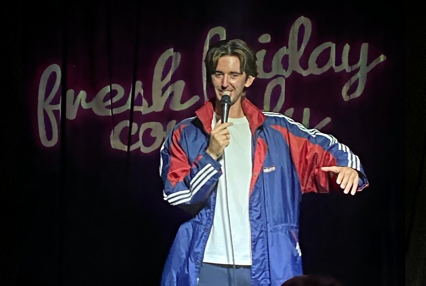 A comedian performing stand-up on a stage, holding a microphone, partly obscuring the words 'fresh friday comedy'  that are projected onto the curtain behind them.