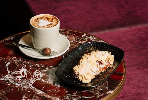 A dark red toned marble table with a coffee and almond croissant on a plate. 