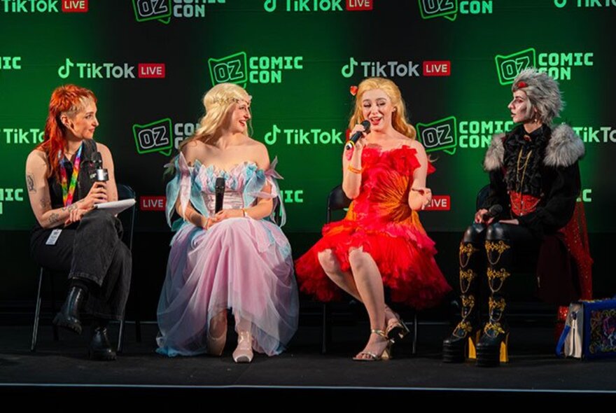 A Comic-Com panel of four women dressed as characters holding microphones and talking on stage. 