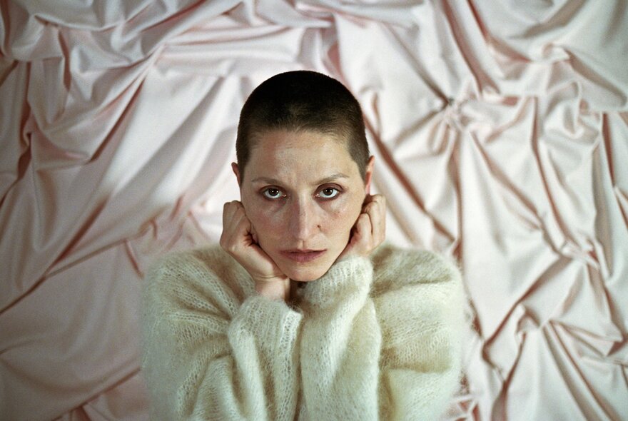 Welsh musician Cate Le Bon wearing a fluffy white jumper, holding her clenched fists up to her cheeks, gazing intently at camera, she has a shaved head and is standing in front of pale pink draped fabric.