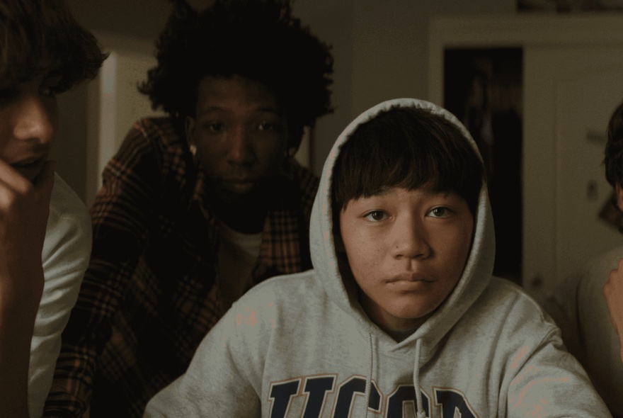 A still shot from the film, Dìdi, with a Taiwanese American boy wearing a grey hoodie with the hood on with two friends behind him, all looking at the camera as though it's a computer screen.