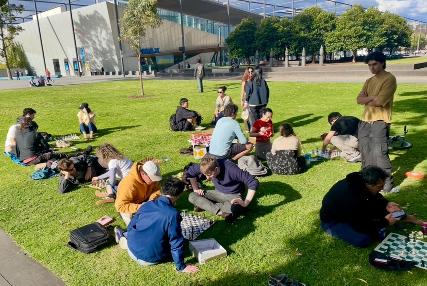 A group of people sitting on the lawns in front of Melbourne Museum playing chess.