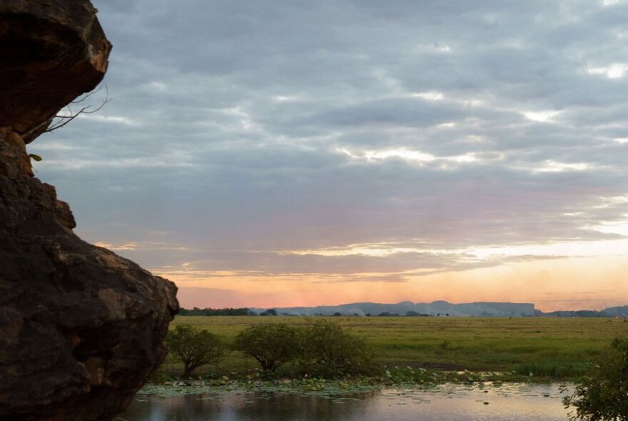 A rocky outcrop overlooks a wide green flood plain under a cloudy sky during sunset.