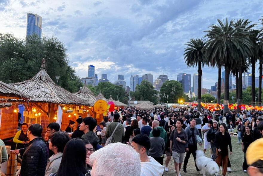 Thatch-roofed market stalls and crowds under palm trees at dusk, with city buildings in the background.