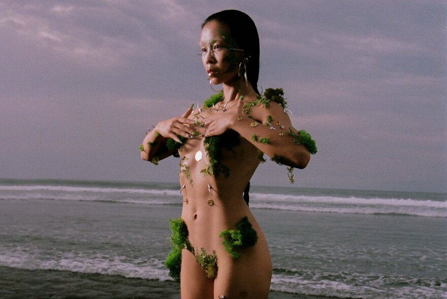 An asian woman standing on a beach wearing only patches of seaweed.
