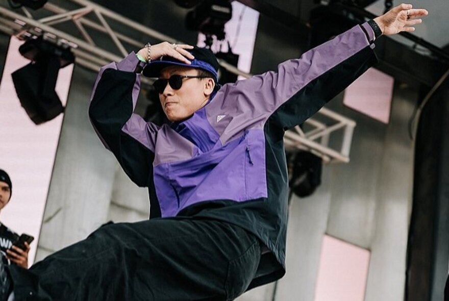 A breakdancer wearing a purple jacket, hat and sunglasses, dancing on a stage.