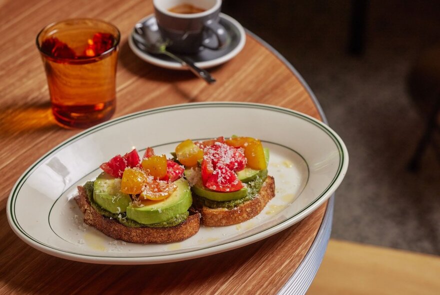 A serve of avocado on toast with toppings next to a glass of water and espresso in a cup.