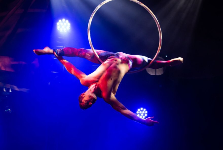 An acrobatic performer hanging upside down in mid air from a hoop, their legs split.