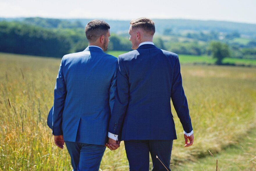 Two grooms wearing blue suits walking hand in hand though an open field, seen from behind.