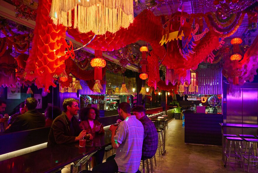 Four people drinking in a neon-lit bar with red Chinese decor hanging from the ceiling.